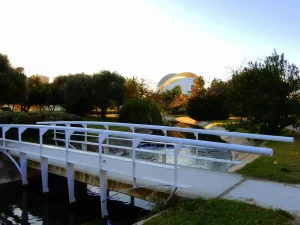 A modern white pedestrian bridge with tubular railings crosses a narrow waterway in Turia Garden. In the background, the distinctive architecture of the Palau de les Arts Reina Sofia is visible amidst trees.