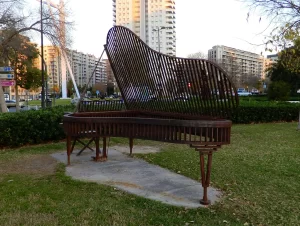 A large, artistic metal sculpture of a grand piano sits on a concrete slab in the grassy Turia Garden, with apartment buildings visible in the background