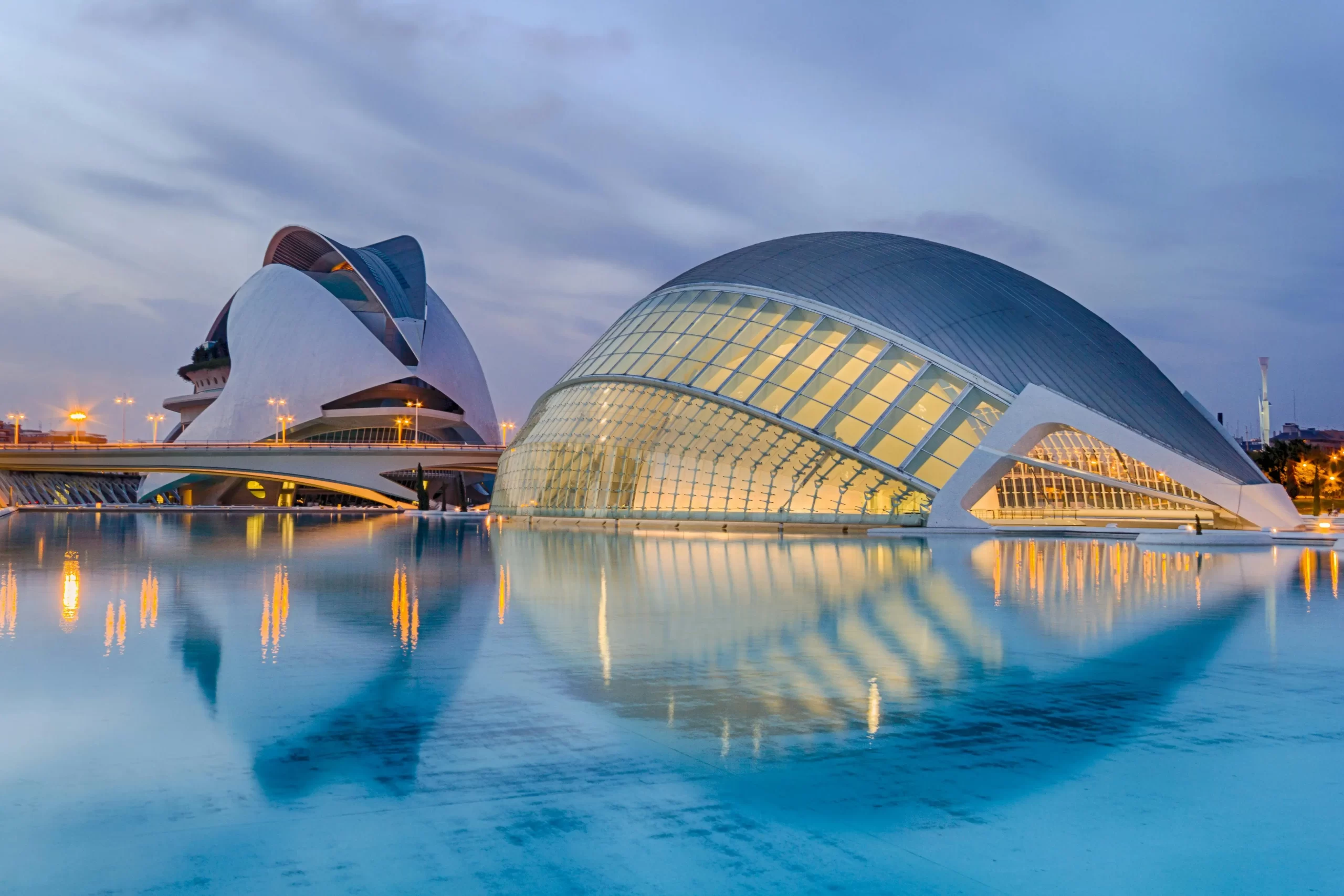Valencia's City of Arts and Sciences complex, featuring L'Hemisfèric and Palau de les Arts Reina Sofía, illuminated at twilight with reflections in the calm water.