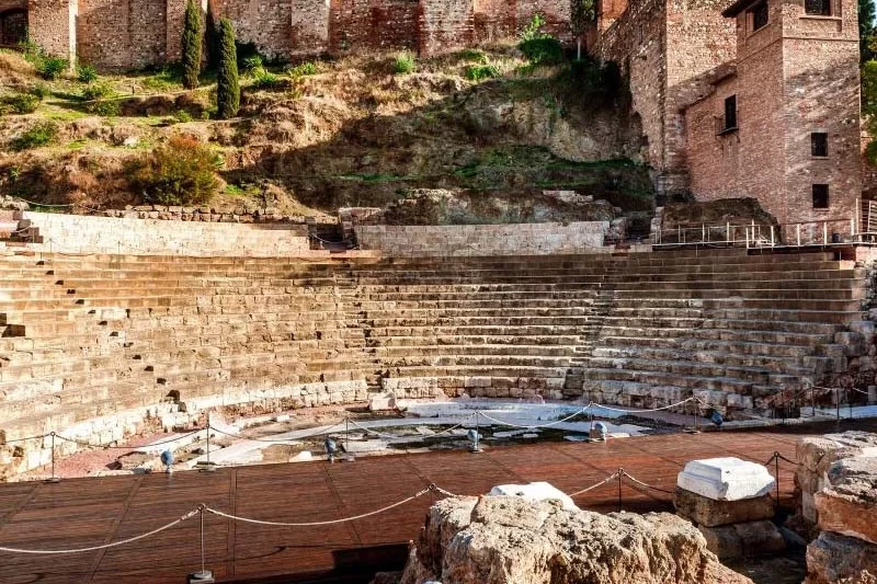 View of the ancient Roman Theatre in Málaga, with its tiered stone seating, situated at the base of a hill.