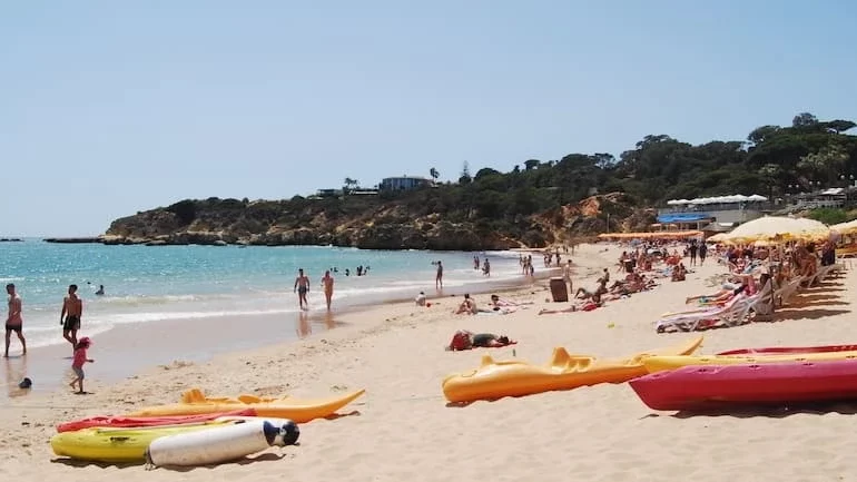 A sunny day at Oura Beach in Albufeira, with people swimming in the turquoise water and relaxing on the golden sand among colorful kayaks.